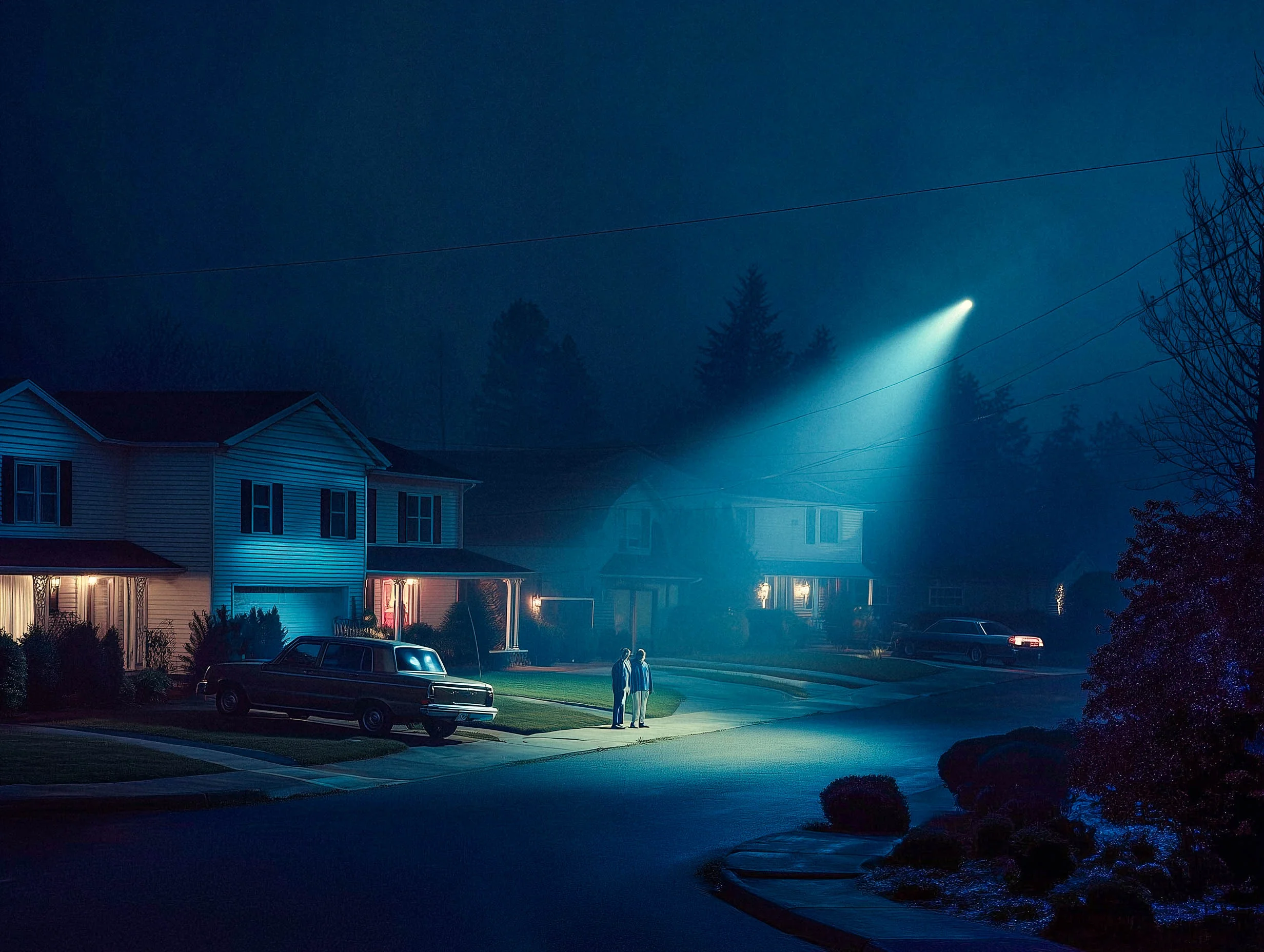 Wide diffused beam on a suburban street at night, fog catching light, two figures, the whole street visible